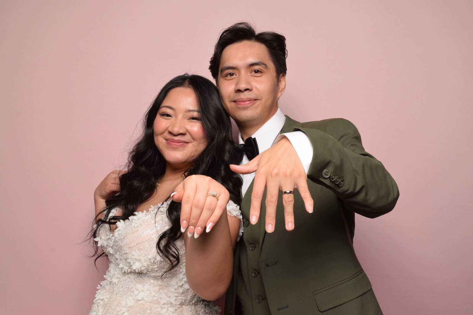 Newlyweds showing their wedding rings against a pink photo booth backdrop